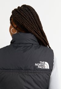 Kobieta z długimi zaplecionymi włosami i srebrną kolczykiem kołowym nosi czarną kamizelkę bez rękawów marki The North Face na jasnej koszuli.