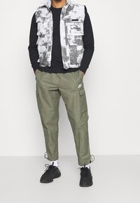 Grå och vit tie-dye-väst med flerfack, svart långärmad tröja, olivgröna cargo-byxor med sidofickor, svarta sneakers.