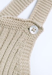 Bretelle beige in maglia con costine testurizzate e un bottone rotondo a quattro fori in madreperla; filato morbido con dettagli di cucitura visibili.