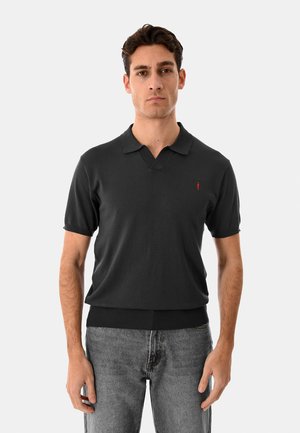 Uomo che indossa una polo nera a maniche corte con piccolo logo rosso sul petto e jeans grigi sbiaditi, in piedi davanti a uno sfondo chiaro e semplice.