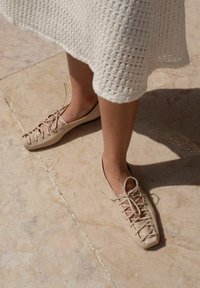 Beige ploché boty se tkaným šněrováním, vyrobené z hladkého materiálu; nošeny s texturovanými bílými pletenými šaty na světlém kamenném povrchu.