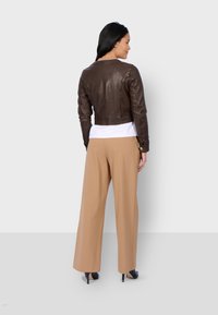 Giacca cropped in pelle marrone, abbinata a pantaloni beige a gamba larga e tacchi alti neri. Presenta dettagli di cucitura e polsini risvoltati.