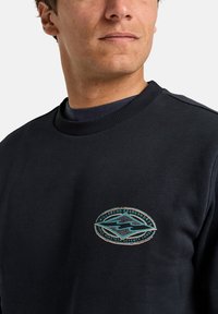 Dark navy sweatshirt with ribbed neckline, featuring a circular embroidered logo in turquoise and white, depicting waves and text details.