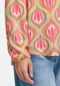 Blouse à manches longues avec un motif floral rétro rose, jaune et vert, associée à un jean en denim bleu clair, main détendue sur le côté.
