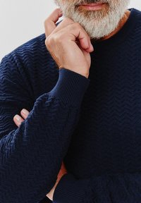 Granatowy sweter dzianinowy o wzorze w rybią ość, z wykończonymi ściągaczami mankietami i okrągłym dekoltem, zaprezentowany na dłoni opierającej się na brodzie.