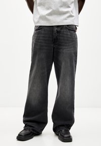 Jeans negros de pierna ancha, fabricados en denim con un lavado claro. Presentan un diseño clásico de cinco bolsillos y costuras visibles. Llevados con calzado negro.