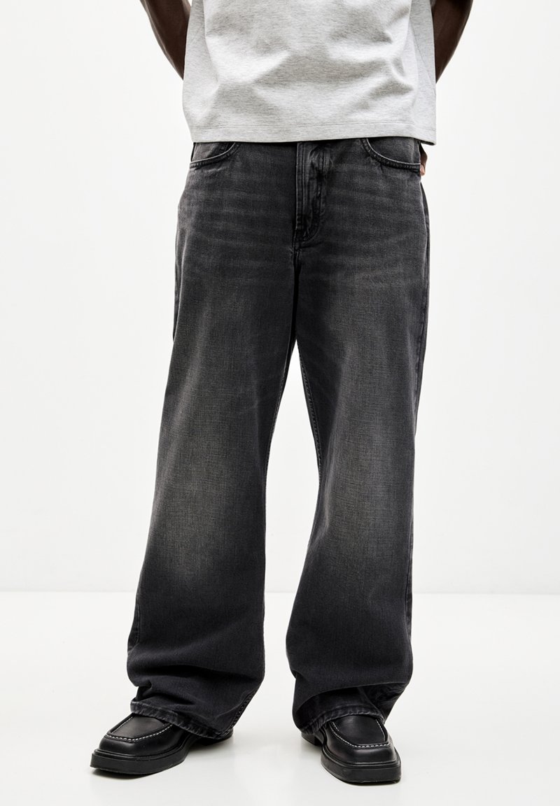 Jeans negros de pierna ancha, fabricados en denim con un lavado claro. Presentan un diseño clásico de cinco bolsillos y costuras visibles. Llevados con calzado negro.