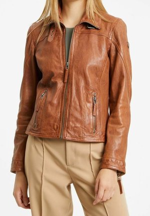 Kobieta w dopasowanej brązowej skórzanej kurtce z zamkiem z przodu i kieszeniami, w połączeniu z beżowymi spodniami i zielonym topem pod spodem.