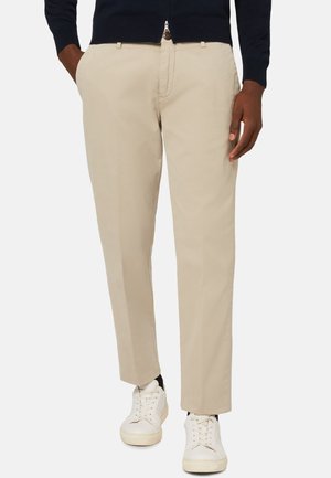 Beige Baumwollhosen mit geradem Schnitt, ausgestattet mit einem Reißverschluss und seitlichen Taschen, kombiniert mit weißen Sneakers. Glatte Textur.