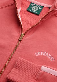 Chaqueta coral con cremallera dorada y puños acanalados. Texto bordado "SUPERDAY" en blanco y acabado interior texturizado.