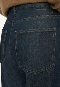 Gros plan sur l'arrière d'un jean en denim bleu foncé avec des coutures visibles et des poches carrées, porté avec un pull marron rentré.