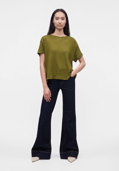 Femme debout, portant un t-shirt ample vert olive, un jean évasé foncé et des chaussures pointues beiges sur un fond blanc uni.
