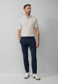 Hellgraues Polohemd mit Kragen, marineblaue Chinohose und weiße Sneakers mit beigen Akzenten, vor einem neutralen Hintergrund stehend.