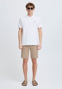 Witte poloshirt, beige shorts en bruine sandalen. De polo heeft een kraag en een klein logo op de borst. Het model draagt een zonnebril met donkere glazen.