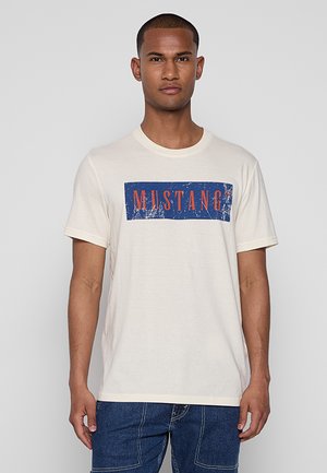 T-shirt en coton beige avec un graphisme rectangulaire comportant "MUSTANG" imprimé en rouge sur un fond bleu. Design à col rond, manches courtes.