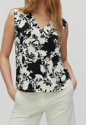Mujer con top negro y blanco sin mangas con estampado floral y escote en V y pantalones claros, de pie con las manos en los bolsillos.