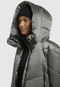 Puffjacka i metallgrått med en stor huva, som har tryckknappar, en ficka med dragkedja och en slät, glänsande yta.