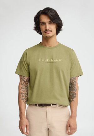 Mężczyzna o średniej długości ciemnych włosach i tatuażach na obu przedramionach ma na sobie oliwkowozieloną koszulkę "Polo Club" i beżowe spodnie.