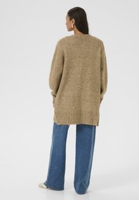 Beżowy sweter dzianinowy o luźnym kroju, z prążkowanym dołem i opadłymi ramionami, w połączeniu z niebieskimi spodniami typu wide-leg, widok z tyłu.