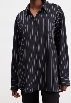 Chemise noire à manches longues avec des rayures verticales blanches et une poche poitrine portée par une personne.
