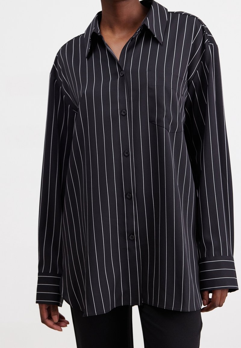 Chemise noire à manches longues avec des rayures verticales blanches et une poche poitrine portée par une personne.