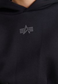 Črna kapuca z mehkim, teksturiranim materialom, ki ima nevsiljivo sivo logotip in tri vodoravne črte. Oblikovanje se osredotoča na minimalizem in udobje.