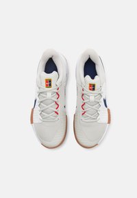 Light gray athletic shoes with a textured upper, navy accents, red laces, and a gum rubber sole. Features a logo on the tongue.