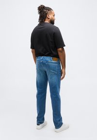 Άνδρας με ντραντλοκς, φορώντας μαύρο μπλουζάκι, μπλε τζιν και λευκά αθλητικά παπούτσια, στέκεται γυρισμένος με την πλάτη σε απλό ανοιχτό φόντο.