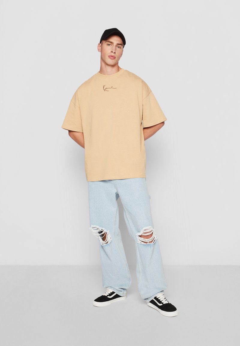Béžové oversized tričko s malým hnedým logom, skombinované so svetlomodrými roztrhnutými džínsami a čiernymi teniskami s bielou podrážkou. Čierna čiapka.