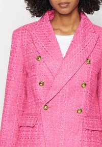 Femme portant un blazer croisé texturé rose vif avec six boutons dorés et poches à rabat sur un top blanc.