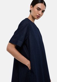 Robe en denim bleu marine, à la coupe décontractée, avec des manches courtes, un col rond et des poches latérales. Tissu lisse et texturé avec des coutures surpiquées.