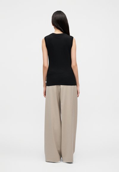 Γυναίκα με μακριά σκουρόχρωμα μαλλιά στέκεται πλάτη, φοράει αμάνικο μαύρο τοπ και φαρδιά μπεζ παντελόνια μπροστά σε απλό φόντο.