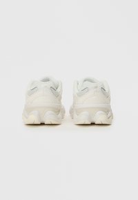 Biele tenisky s textúrovaným zvrškom, sieťovanými akcentmi a dizajnom podošvy pripomínajúcim vlnu. Majú polstrovanie okolo goliera a minimálne branding.