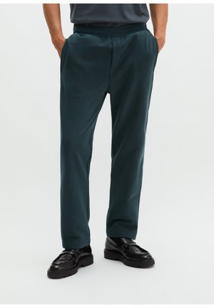 Ein Mann trägt dunkelgrüne Hosen mit Seitentaschen und schwarze Lederschuhe und steht vor einem schlichten hellen Hintergrund.