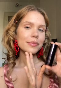 Tube de maquillage cylindrique noir tenu dans une main, avec une personne portant un haut à carreaux et des boucles d'oreilles en forme de fleurs rouges. Les cheveux sont ondulés et lâches.