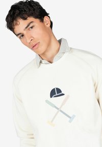 Sudadera color marfil con un suave diseño bordado de un casco de equitación y dos palos de polo cruzados en colores pastel.