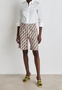 Henkilö, joka on pukeutunut valkoiseen nappipaitaan, vaaleanpunaisiin ananaskuvioisiin shortseihin ja keltaisiin korkokenkiin, seisoo vaalealla lattialla.