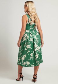 Vihreä kukkakuosis midi-mekko, jossa on valkoisia kukkia, istuva yläosa, pleatattu takasuunnittelu ja yhdistetty mustiin korkokenkiin.