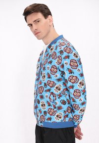 Jachetă albastră tip bomber, cu modele complexe de cranii de zahăr și flori, sistem de închidere cu fermoar și manșete ribbate, cu o textură ușoară.