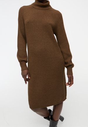Femme portant une robe à col roule marron en maille côtelée avec manches longues et des bottines noires, debout devant un fond blanc uni.