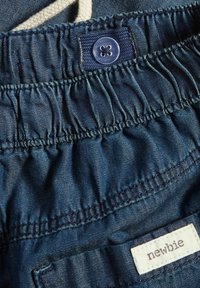 Farkkushortsit, joissa on joustava vyötärö, vaaleansininen väri, röyhelöinen pinta, tummalla napilla ja kankaalla varustettu etiketti, jossa lukee "newbie".