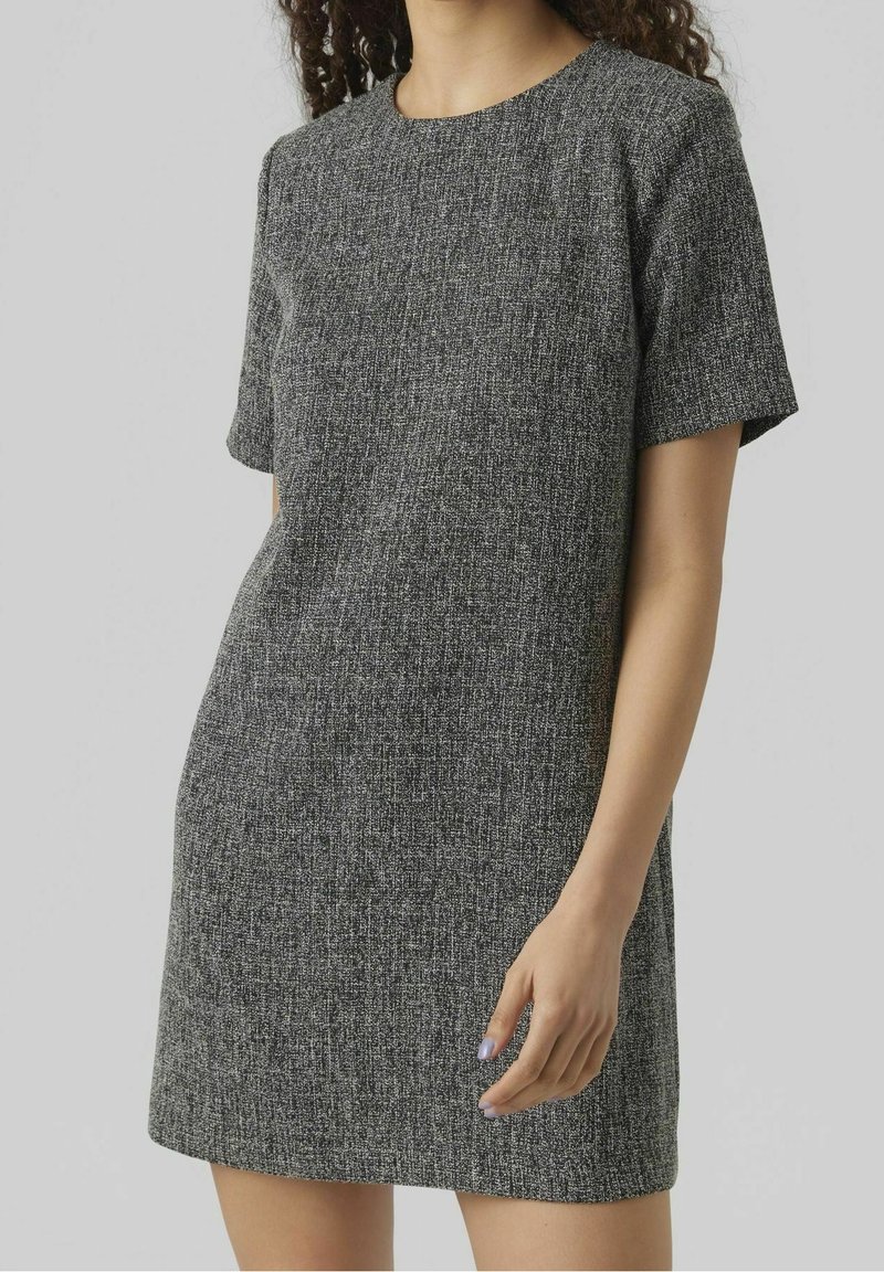 Femme portant une robe grise texturée à manches courtes et longueur genou, avec un col rond, debout devant un fond clair uni.