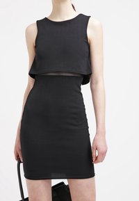 Vestido negro sin mangas con silueta ajustada, detalle de top corto, textura suave y largo a mitad del muslo, llevado con un bolso negro.
