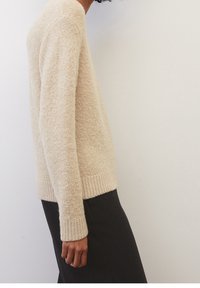 Pull en maille beige avec une finition douce et texturée, muni de poignets et d'un ourlet côtelés. Porté avec un pantalon foncé, montrant une coupe décontractée.