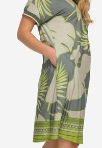 Szara sukienka w zielone tropikalne wzory liści. Posiada krótkie rękawy, kieszeń oraz geometryczne wykończenie u dołu. Lekka tekstura materiału.