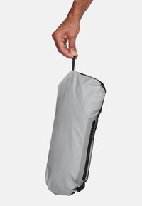 Hand houdt een grijs, compact etui met ritssluiting vast aan een klein zwart stoffen lusje tegen een effen achtergrond.