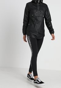 Svart vattentät jacka med frontficka, justerbar huva och elastiska ärmar, kombinerad med svarta leggings som har en vit mönstrad remsa. Svarta sneakers.