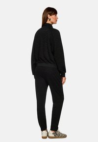 Schwarzer, strukturierter Trainingsanzug mit einem dezenten Muster, ausgestattet mit einem hohen Kragen und elastischen Bündchen. Kombiniert mit gemusterten Sneakers.