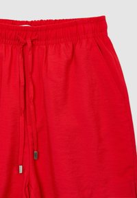 Short en tissu rouge avec une taille élastique, des cordons à embouts métalliques et une poche latérale à droite.