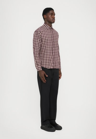 Uomo in piedi, che indossa una camicia a quadri bordeaux e bianca, pantaloni neri e sneakers nere, su uno sfondo bianco semplice.